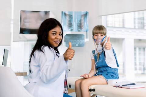 A female nurse and an child are in the medical room and both are giving a thumbs up